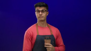 Kırmızı gömlekli ve önlüklü İspanyol genç adam parlak mavi arka planda kahve fincanı tutuyor..