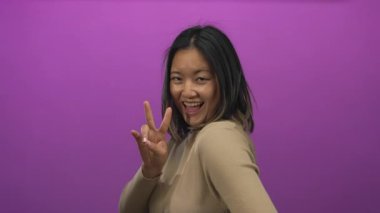 Canlı mor bir duvara karşı barış işaretiyle gülümseyen kadın, elleri havada, bej bir süveter giymiş, renkli bir arkaplanda izole edilmiş, neşeli bir anı yakalıyor..