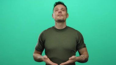 Genç İspanyol adam, izole edilmiş yeşil arka plana karşı meditasyon yapıyor. Sakince ifadesini ve dövmelerini gösteriyor. Rahatlama ve farkındalık taşıyor.