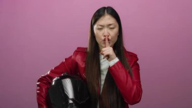 Kırmızı ceketli bir kadın, pembe bir arka planda parmaklarıyla motosiklet kaskı tutuyor. Sessizlik ya da gizlilik öneriyor. İlginç bir şekilde gizemli bir ruh hali içinde..