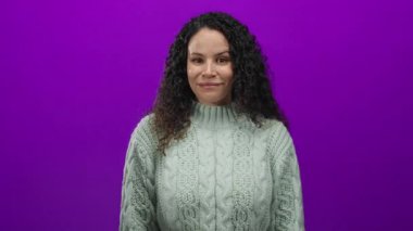 Kıvırcık saçlı, gülümseyen ve göz kırpan canlı mor arka planda rahat bir kazak giyen stüdyo ortamında neşeli bir ruh hali yakalayan bir kadın..