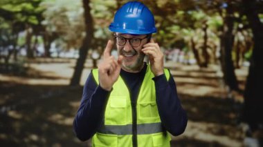 İnşaat malzemesi giymiş olgun bir adam açık hava parkında jestlerle telefonda konuşuyor..