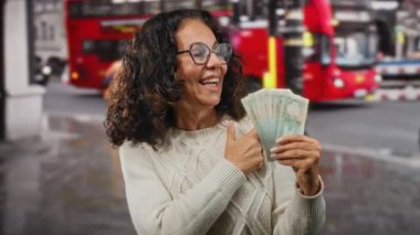 Başparmakları havada gülümseyen bir kadın, arka planda ikonik kırmızı otobüsler olan hareketli bir caddede, hareketli bir şehir ortamını yakalayan kirli para banknotlarını tutuyor..