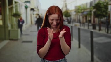 Kızıl saçlı bir kadın canlı bir şehir caddesinde alkışlıyor ve gülümsüyor, arka planda insanlarla birlikte açık bir şehir ortamında neşe ve enerji sergiliyor..