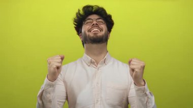 Başarıyı kutlayan genç adam gözleri kapalı ve yumrukları parlak sarı bir arkaplanda, mutluluk ve zaferi ifade ediyor..