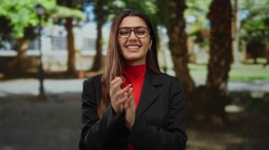 Güneşli bir park ortamında mutlu bir şekilde alkışlayan, gözlük takan, canlı kırmızı ve siyah kıyafetli bir kadın, dışarıda olumlu ve başarılı olma hissi uyandırıyor..