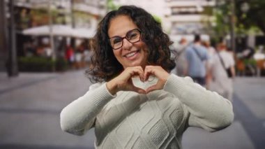 Kıvırcık saçlı, gülümseyen bir kadın kalp şeklinde, şehir sokaklarında elleri açık, süveter ve gözlük takan, mutluluk ve aşkı gösteren bir kalp..