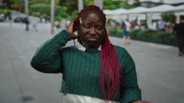 Pembe örgülü yeşil kazaklı bir kadın şehir caddesinde dikiliyor. İnsanlar bulanık arka planda yürürken belirsiz görünüyor..