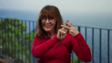 Kırmızı kazaklı İspanyol kadın hashtag jesti yapıyor. Okyanusun kenarında manzaralı bir arka plan gibi mutlu bir şekilde duruyor..