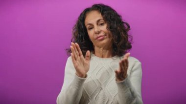 Kıvırcık saçlı bir kadın canlı mor bir arka plana el çırpıyor olgun bir zarafet ve kendinden emin bir ifade sergiliyor ve neşeli bir takdir görüntüsü yaratıyor..