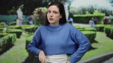 Mavi kazaklı esmer kadın, yemyeşil ve bulanık çit labirenti ile güneşli parkta mizahi bir şekilde el kol hareketi yapıyor, neşeli bir etkileşim ve açık hava atmosferi öneriyor..