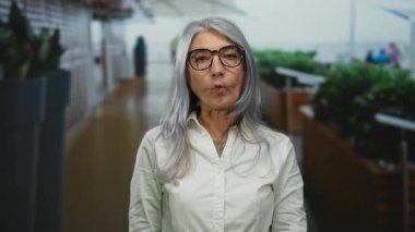 Gri saçlı yaşlı bir kadın açık havada komik bir yüz yapıyor. Etrafı bulanık insanlar ve yeşilliklerle çevrili, gözlüklü ve beyaz gömlekli..