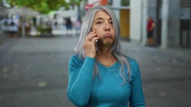 Gri saçlı yaşlı kadın şehir sokaklarında telefonla konuşuyor. İnsanlar ve binalar arasında düşünceli bir yüz ifadesi sergiliyor.