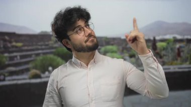 Sakallı İspanyol genç adam yeşil ortamda iyimserlik ve düşünceli düşünme önererek açık hava parkında başparmağını yukarı kaldırıyor ve puan veriyor..