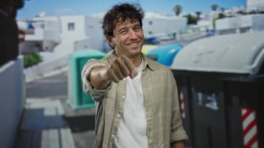 Gülümseyen İspanyol adam, açık mavi gökyüzünün altında kentsel mimari ve geri dönüşüm kutularıyla çevrili bir sokakta kameraya doğru elini uzattı..