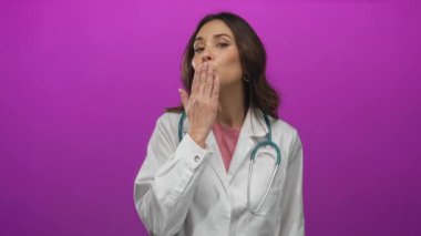 Steteskoplu beyaz önlüklü İspanyol kadın doktor, izole edilmiş pembe arka plana öpücük yolluyor, sağlık hizmetlerinde sıcaklık ve profesyonelliği somutlaştırıyor..