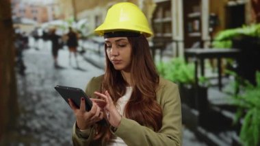 Sarı kask takan ve tablet kullanan bir kadın, açık bir sokakta duruyor, şehir ortamında modern bir mimar ya da mühendis gibi projeler yönetiyor..
