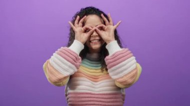 Renkli süveter giymiş, gülümseyen bir kadın. Elleriyle poz veriyor, gözlük takıyor, mor bir arka plana yaslanıyor ve neşe ve yaratıcılık ifade ediyor..