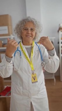 Gri saçlı, hayır kurumunda gönüllü olarak çalışan, steteskop ve rozet takan yaşlı bir kadın, kapalı bir odada kutular ve giysilerle çevrili jestler yapıyor..