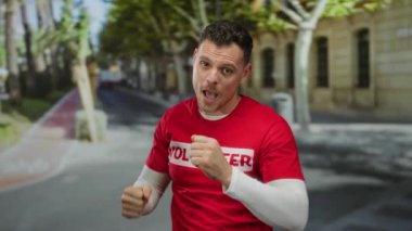 İspanyol bir adam kırmızı gönüllü tişörtü giymiş, arka planda ağaçlar ve binalarla dolu canlı bir şehir atmosferini taşıyan bir şehir caddesinde neşeyle dans ediyor..