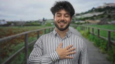 Sakallı genç adam yeşil bir parkta duruyor. Gülümsüyor ve göğsüne dokunuyor. Bulutlu bir gökyüzünün altında huzurlu doğal ortamın tadını çıkarıyor..