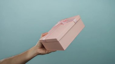 Mavi arka planda pembe hediye kutusunu tutan adamın eli basit bir zarafetle izole edilmiş bir ortamda hediyesini gösteriyor..