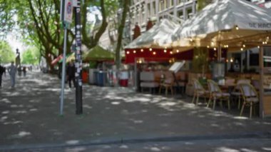 Avrupai bir sokak manzarası bulanık. Açık hava kafeleri ve geçmekte olan insanlar hareketli bir şehir merkezinde yapraklı ağaçların altında çözülmüş bir bokeh etkisi yaratıyor..