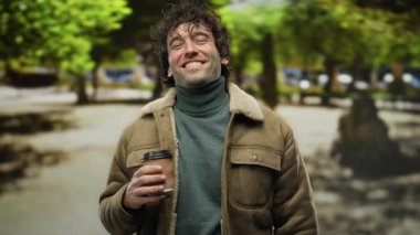 Dışarıda gülümseyen ve elinde bir kahve fincanı tutarken kameraya öpücük yollayan genç adam, arka planda ağaçlar olan bir şehir caddesinde gündelik bir anın tadını çıkarıyor..