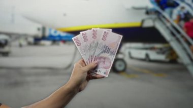 Bir havaalanı terminalinde elinde Türk lira banknotları tutan adamın arka planda bir uçak, seyahat, finans ve uluslararası seyahatin altını çiziyor..