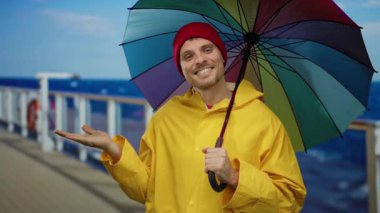 Sarı yağmurluklu genç adam deniz kıyısındaki bir yolcu gemisinde renkli bir şemsiye tutuyor. Mavi gökyüzünün altında kırmızı bir bere takıyor..