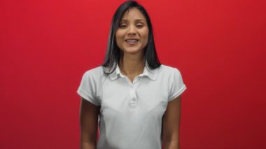 Beyaz tişörtlü, gülümseyen genç İspanyol kadın elleri ile canlı kırmızı arka plana karşı kalp hareketi yapıyor, pozitiflik ve şefkat gösteriyor..