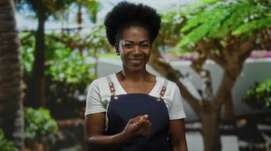 Önlüklü kadın, yemyeşil bir parkta parmaklarıyla bir jest yapıyor. Etrafı yeşillik ve sıcaklıkla çevrili ve gülümsüyor..