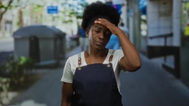 Önlük giymiş bir kadın, şehir caddesindeki açık alanda düşünceli bir şekilde duruyor..