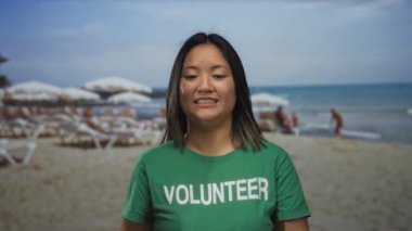 Sahilde yeşil t-shirt giyen genç bir kadın ve arka planda başkaları var. Kum ve okyanus dalgalarına karşı çeşitli duygular ifade ediyorlar..