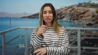 Çizgili kazaklı genç sarışın kadın açık havada okyanus manzaralı bir sahil yürüyüşünde duygularını ifade ediyor ve arka planda kayalık uçurumlar açık bir gökyüzünün altında..