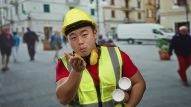 İnşaat malzemesi giymiş genç Çinli adam planlar tutuyor ve hareketli bir sokak ortamında öpücükler saçıyor..