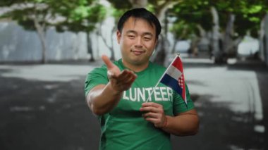 Hırvat bayrağı ve yeşil gönüllü tişörtlü genç adam, arka planda ağaçlarla kent caddesinde gülümsüyor.