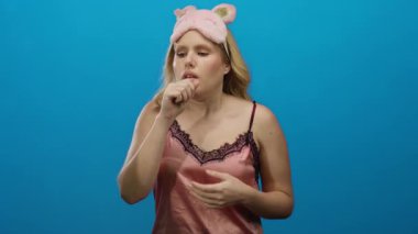 Sarı saçlı, pembe uyku maskesi takan ve mavi arka planda öksüren genç bir kadın.