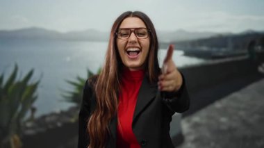 Gözlüklü gülümseyen bir kadın, güneşli deniz kıyısında arka planda dağlar olan kameraya doğru uzanır. Bu da sıcaklık ve açıklığı gösterir..