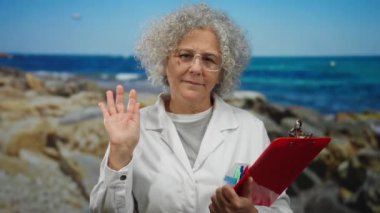 Beyaz önlüklü, kıvırcık gri saçlı kadın, arka planda mavi deniz olan kayalık bir sahilde dururken kırmızı bir pano ve dalgalar tutuyor..