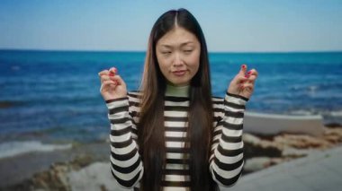 Uzun saçlı bir kadın güneşli bir günde deniz kenarında çizgili gömlek giyerek jest yapıyor.
