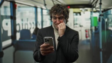 Otobüsteki adam gergin bir şekilde telefona bakıyor. Kıvırcık saçlı ve siyah takım elbiseli. Toplu taşıma araçlarında stres belirtileri var.