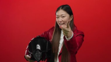 Deri ceket giyen, modern tarzı ve kendine güveni olan, neşeli bir miğferi olan, kırmızı arka planda duran bir kadını işaret ediyor.