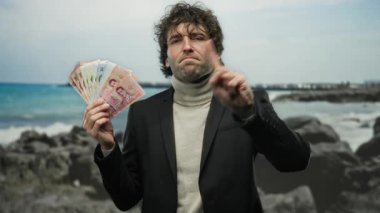 Deniz kıyısındaki İspanyol adam Tayland banknotlarını ciddi bir ifadeyle tutuyor. Onaylamadığını işaret ediyor. Tropikal ortamda finansal tema sergiliyor..
