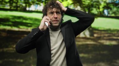 Parktaki genç adam yüzünde endişeli bir ifadeyle, güneşli bir günde siyah bir ceket giyerken cep telefonuyla konuşuyor..