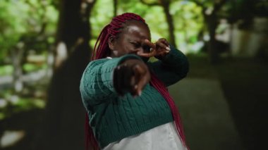 Canlı yeşil bir parkta parmaklarıyla neşeli poz veren bir kadın, yeşil bir kazak giyiyor ve güneşli bir günde neşeli duygular sergiliyor..