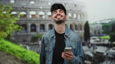Roma Kolezyumu 'nun yanında akıllı telefonu tutarken gülümseyen İspanyol genç adam, şehirde yeşillik olan bir sokak dekorunu gözler önüne seriyor..