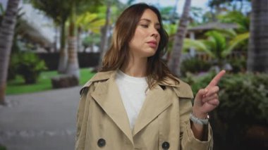 Güneşli parkta bej ceketli bir kadın açık havada yemyeşil bir zemin üzerinde palmiye ağaçlarıyla rahatlamış atmosferi yakalıyor..