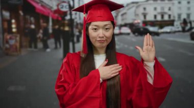 Kırmızı mezuniyet elbisesi giymiş bir kadın, şehir caddesinde elleri yüreğinde duruyor ve şehir binaları ve insanlarla çevrili bir şekilde selamlanıyor..