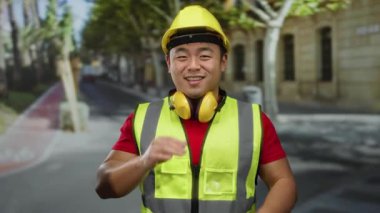 Şehir caddesinde bağıran, güvenlik yeleği ve şapka takan Asyalı genç adam, şehir dışında inşaat sahnesini gösteriyor, hareketli bir ortamda iletişim enerjisi ve nişan taşıyor..
